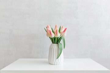 Light pink tulips flowers in a white geometric ceramic vase stand on a white table near grey wall.