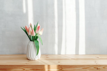 Light pink tulips in a white geometric ceramic vase stand on a wooden table near grey wall. Bouquet of flowers in the morning sun beams.