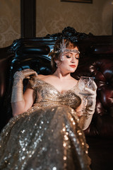 a girl in a gold dress with a diadem on her head, sits on a leather chair, with one hand shows her...