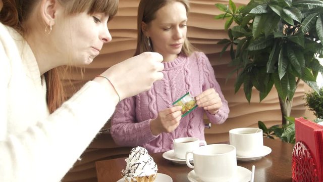 Two pretty woman friends are conversating news, drinking coffee and eating ice cream on coffee break in cafe with decorative tree and big window on background. Closeup portrait view shot 4K video.