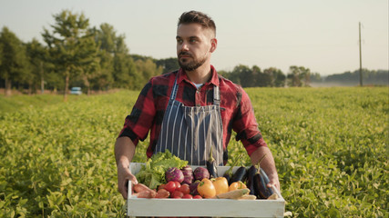 Young handsome farmer is holding a box of organic vegetables look at camera at sunlight agriculture...
