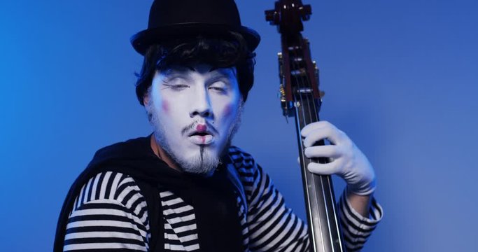 Close up handsome male mime clown emotionally plays on contrabass in black and white striped clothes. Isolated blue background
