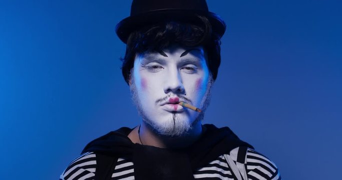 Close up of handsome male mime seriously looking into a camera and smoking cigarette in black and white striped clothes. Isolated blue background