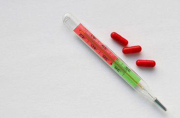 Glass thermometer and three red capsules on a white background. Selective focus. Copy space. Medical supplies and medicines. Treatment and health care