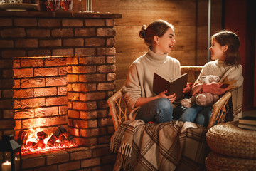 family mother and child reading book and drink tea on winter evening by fireplace