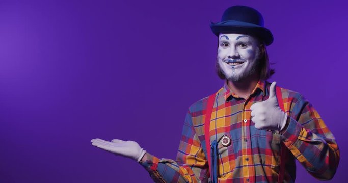 Handsome colorful charismatic smiling mime clown in a hat shows and performs copy space by his hands. Purple isolated background.