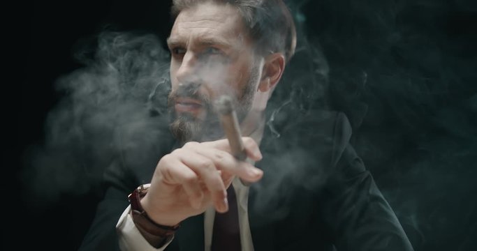Pensive bearded male in elegant suit smoking cigar in dark room full of smoke, closeup
