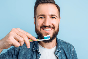A young handsome man with a beard is brushing his teeth on an isolated blue background with raised