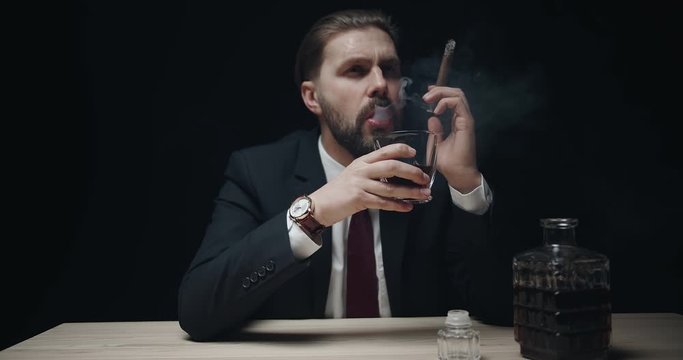 Handsome bearded male in elegant formal suit smoking cigar and drinking whiskey isolated on black background