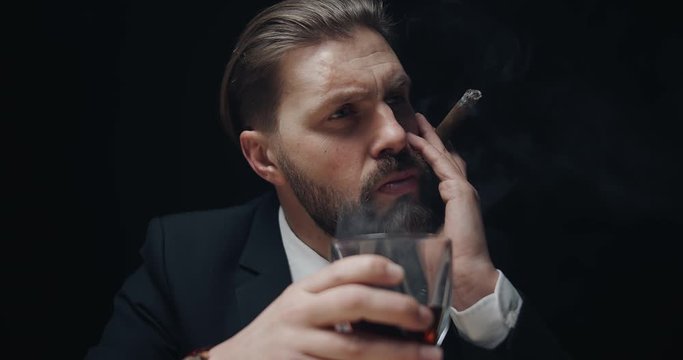 Brutal bearded middle aged male in black suit smoking cigar and drinking whiskey in dark room, gangster