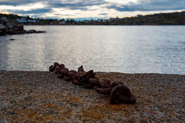 A large rusty chain attached to a breakwater wall in a wide open harbour.  The sky is cloudy and grey with lots of trees and hills. The ocean is calm with small ripples. 