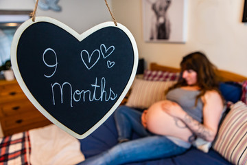 A selective focus shot of a heart chalkboard sign saying 9 months. with a blurry pregnant woman lying in bed resting hands on baby bump in background