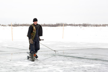 Fototapeta na wymiar A worker walks on the ice of a frozen lake, with a sawing machine