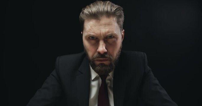 Bearded middle-aged man in elegant black suit looking weirdly to camera isolated on black background, evil look