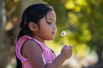 A thouthful little kid holding a dandelion in her hand that is close to her face and surrounded by...