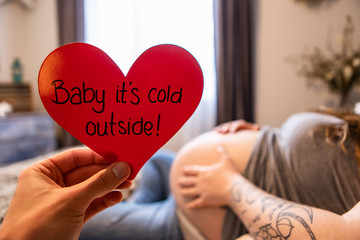 A selective focus first person perspective of a hand holding a red heart with message baby its cold outside, as heavily pregnant woman rests in bed