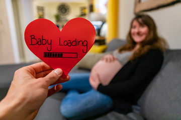 A first person perspective of a father to be holding a red valentine heart with the message baby loading, a heavily pregnant woman is seen blurry in background