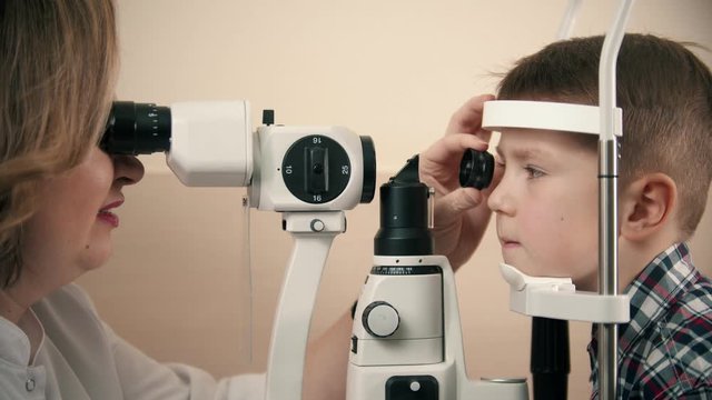 Boy having a treatment in eye clinic - a woman doctor checking little boy's eye vision by looking through big special device using a lens and bright lighting