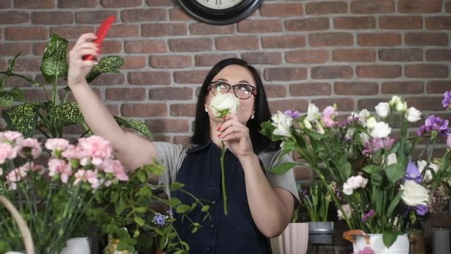 Small private flower business. a professional florist in glasses and apron in a flower shop takes a selfie with a white rose on the phone camera. The girl takes photos for social networks.