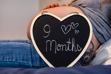 A close up shot of a cute heart shaped sign with handwritten message saying 9 months, resting against the large stomach of a heavily pregnant woman