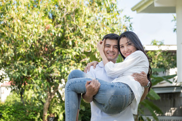 Fototapeta premium Young loving couple have fun in park. Caucasian boyfriend carry on his asian girlfriend in arms in front yard of house.