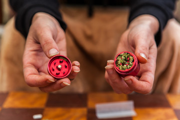 Selective focus closeup of person's hands holding a red weed filled grinder. Cannabis buds inside a grinder, checkered out of focus table in background.