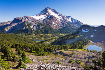 A high elevation volcanic peak seen in daylight in the summer with little snow and ice remaining on the slopes above tree line.