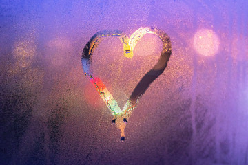 Close-up drawn heart on a foggy transparent window with bright multi-colored blurred city lights in the background. Symbol of love on wet glass