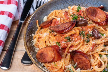 A bowl of chef prepared spaghetti with chorizo, black olives, and spinach in a creamy tomato sauce in a ceramic bowl on a wooden chopping board with red and white napery.