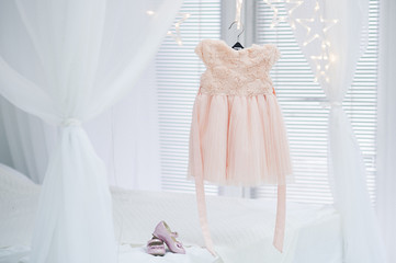 preparation for the holiday for the girl, a delicate pink tulle dress weighs on a hanger against a light background of beds and curtains, pink little shoes stand nearby