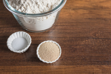 White flour in a glass bowl container, dry yeast, and salt in small ramekin on wood table surface.