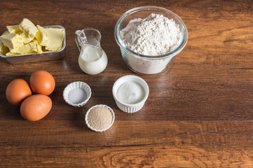 White flour in a glass bowl container, dry yeast, salt, and sugar in small ramekin. 3 pieces of brown chicken eggs, butter, and milk in small glass jar on wood table surface.