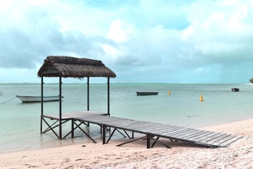 Over water thatched hut, rustic bungalow with a wooden planked walkway installed on a white sand beach.