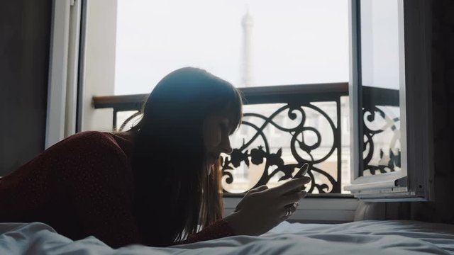 Happy beautiful European woman typing messages on smartphone app lying on balcony bed with Eiffel Tower view slow motion