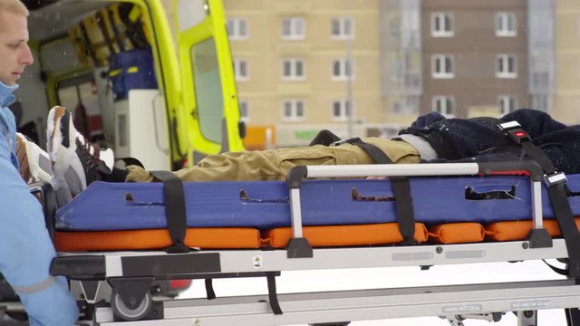 Tracking shot of male paramedics in uniforms loading unconscious patient on stretcher into ambulance on cold winter day