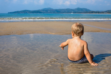 Happy little child sitting in water on seashore in summer back view