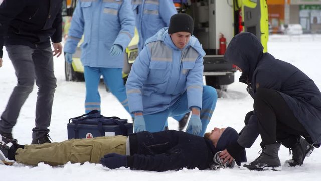 Tracking of worried people sitting beside unconscious man lying on snow as team of paramedics arriving on scene