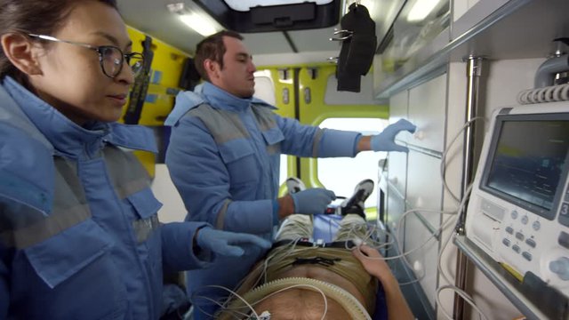 Handheld tracking shot of male and female EMTs talking and monitoring vitals of unconscious patient lying on stretcher inside moving ambulance