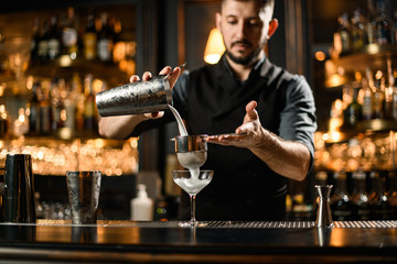Male bartender pouring an alcoholic cocktail drink from the steel shaker through the sieve