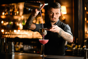 Male bartender pouring a pink alcoholic cocktail drink from the steel shaker through the sieve
