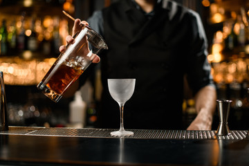 Male bartender pouring a brown alcoholic cocktail drink from the glass through the strainer