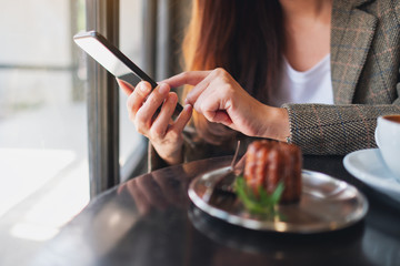 Closeup image of a woman holding and using mobile phone with coffee cup and snack on the table in cafe