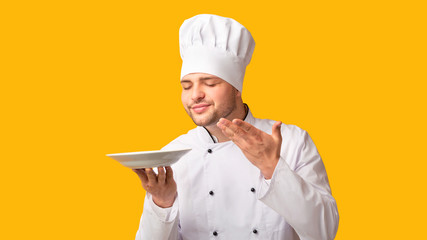 Professional Chef Holding Empty Plate Smelling Invisible Dish, Yellow Background