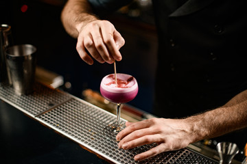 Male bartender stirring a crimson alcoholic cocktail drink in the glass with a toothpick