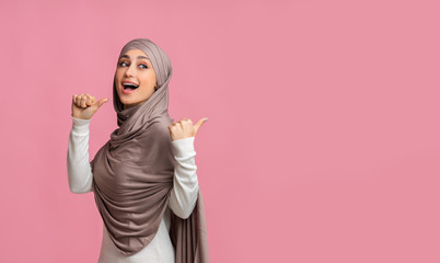 Joyful muslim woman in hijab pointing at copy space behind her
