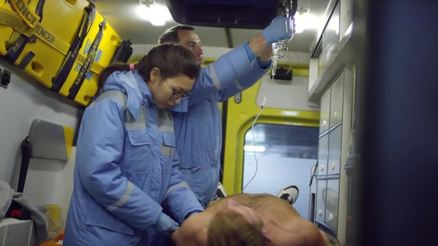 Handheld shot of female paramedic inserting needle into arm of unconscious patient lying in ambulance as male EMT preparing IV bag
