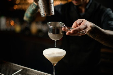 Male bartender pouring a creamy alcoholic cocktail drink from the steel shaker through the sieve