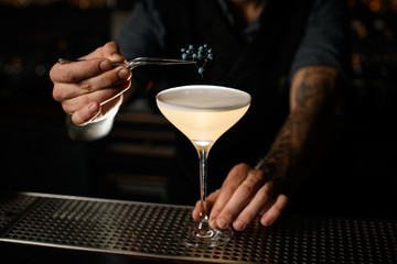 Professional bartender decorating a creamy alcoholic cocktail in the glass with a dried flower by tweezers