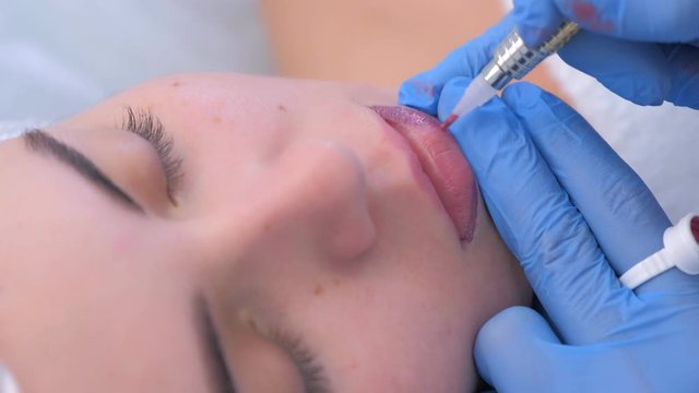 Cosmetologist making lips microblading procedure for girl using tattoo machine and pink pigment. Beautician applying permanent makeup on woman lips. Beauty industry concept. Client's face closeup.