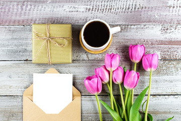 Flat Lay Composition with spring pink tulips flowers, gift box, cup of coffee and empty card on shabby wooden background. Greeting card for Womens or Mothers Day. Top view, copy space. Mockup concept.